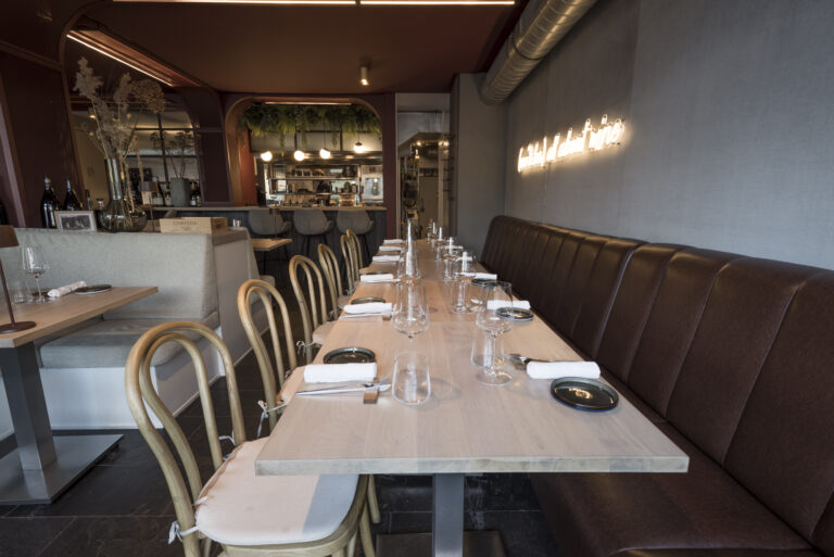Stijlvol restaurant interieur met lange gedekte houten tafel, bruine lederen bank, rotan stoelen en neonverlichting.