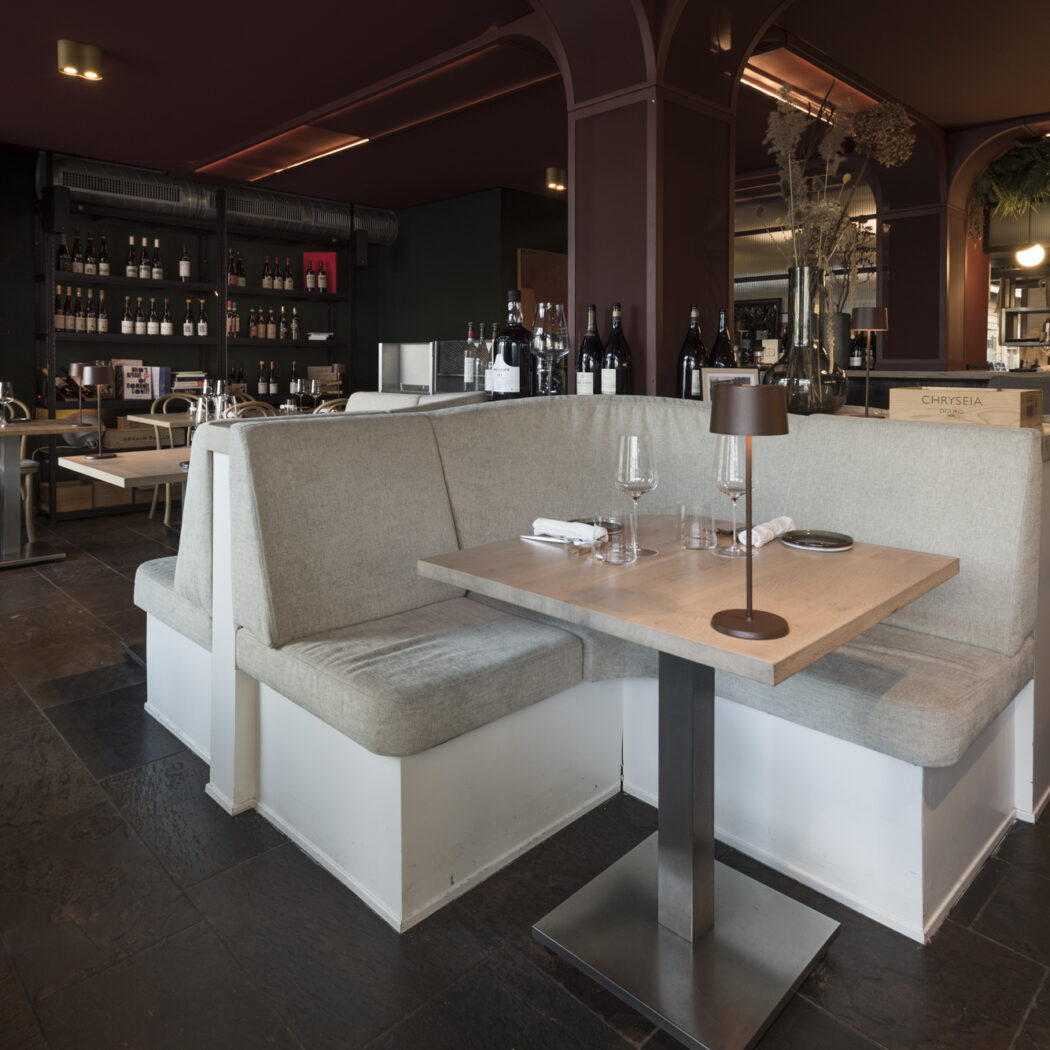 Modern restaurant met grijze hoekbanken, gedekte houten tafels, wijnrekken en een bar met CHRYSEIA kist.