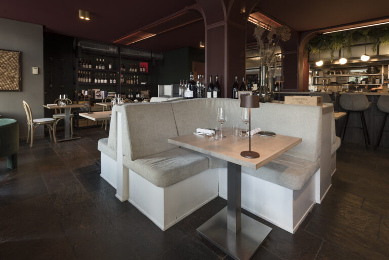 Modern restaurant met grijze hoekbanken, gedekte houten tafels, wijnrekken en een bar met CHRYSEIA kist.