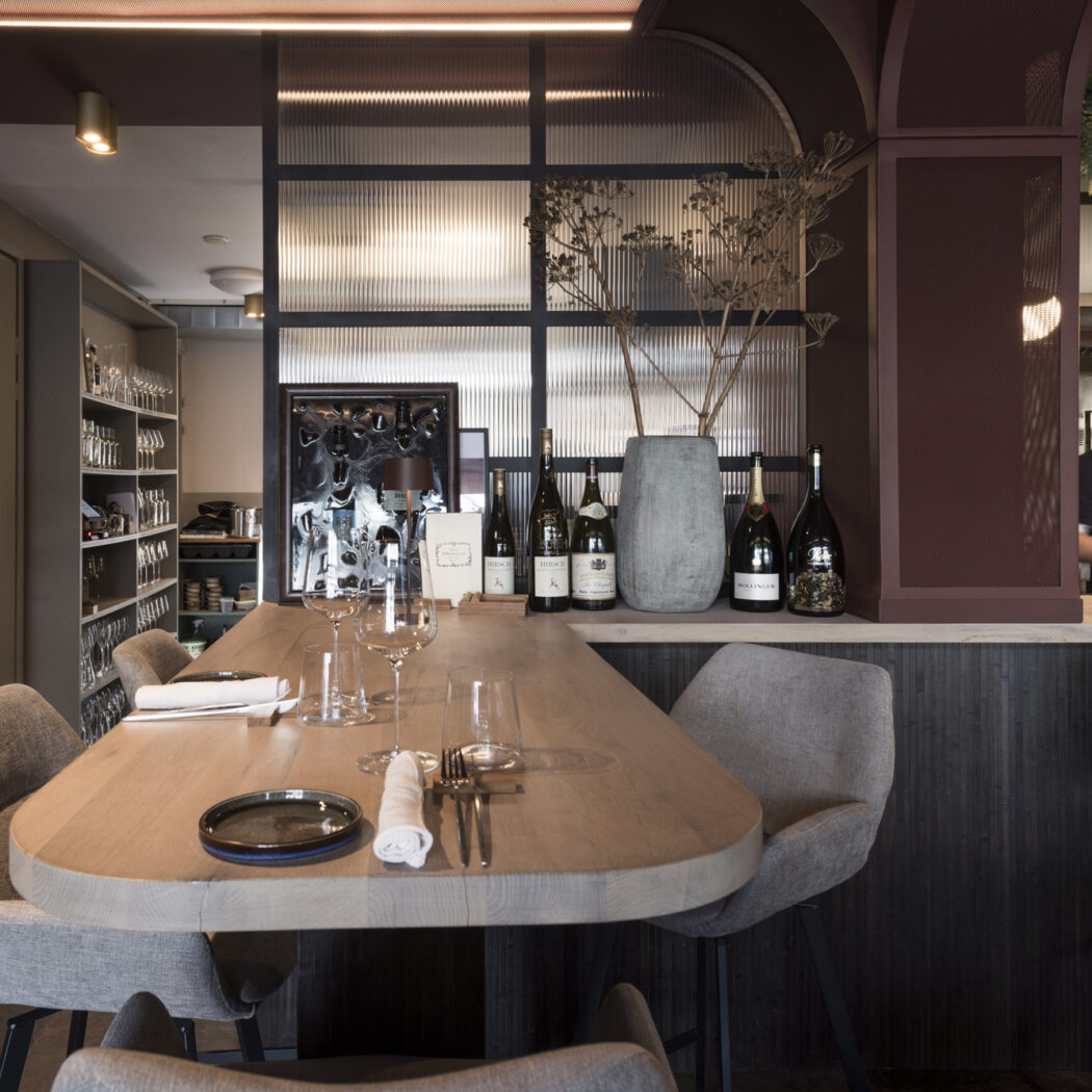 Stijlvol restaurantinterieur met gedekte houten tafel, wijnen op de counter en een medewerker.