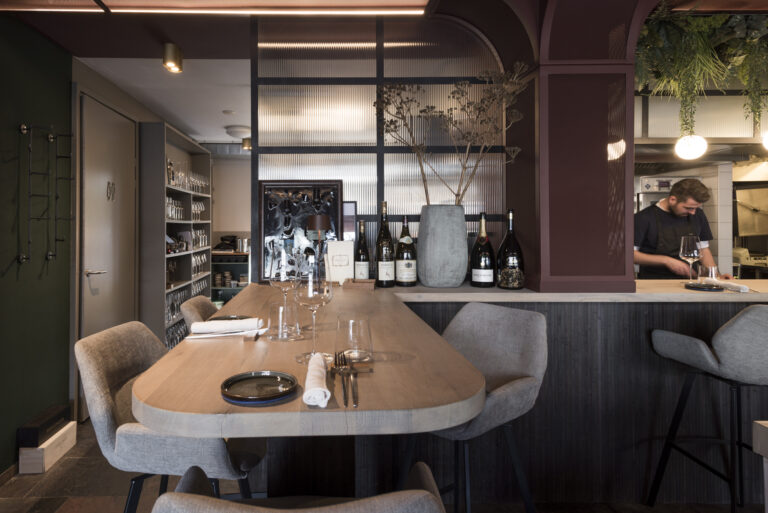 Stijlvol restaurantinterieur met gedekte houten tafel, wijnen op de counter en een medewerker.