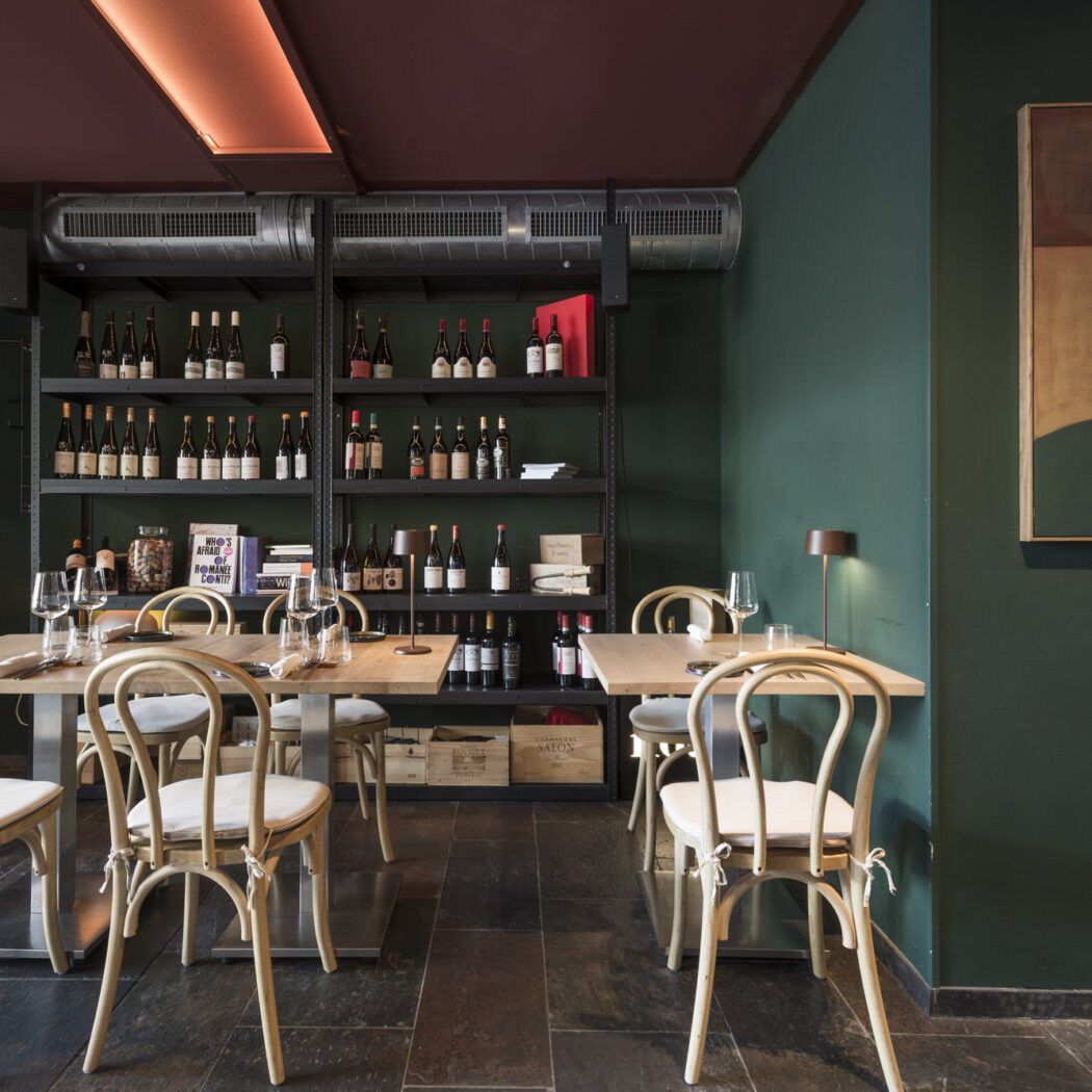 Een sfeervol restaurant met donkergroene muren, houten tafels en stoelen, en rekken vol wijnflessen.