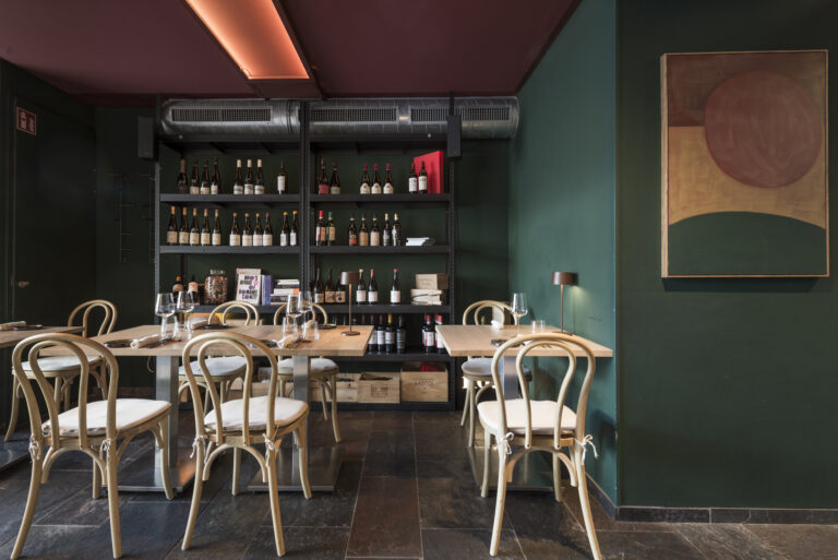 Een sfeervol restaurant met donkergroene muren, houten tafels en stoelen, en rekken vol wijnflessen.