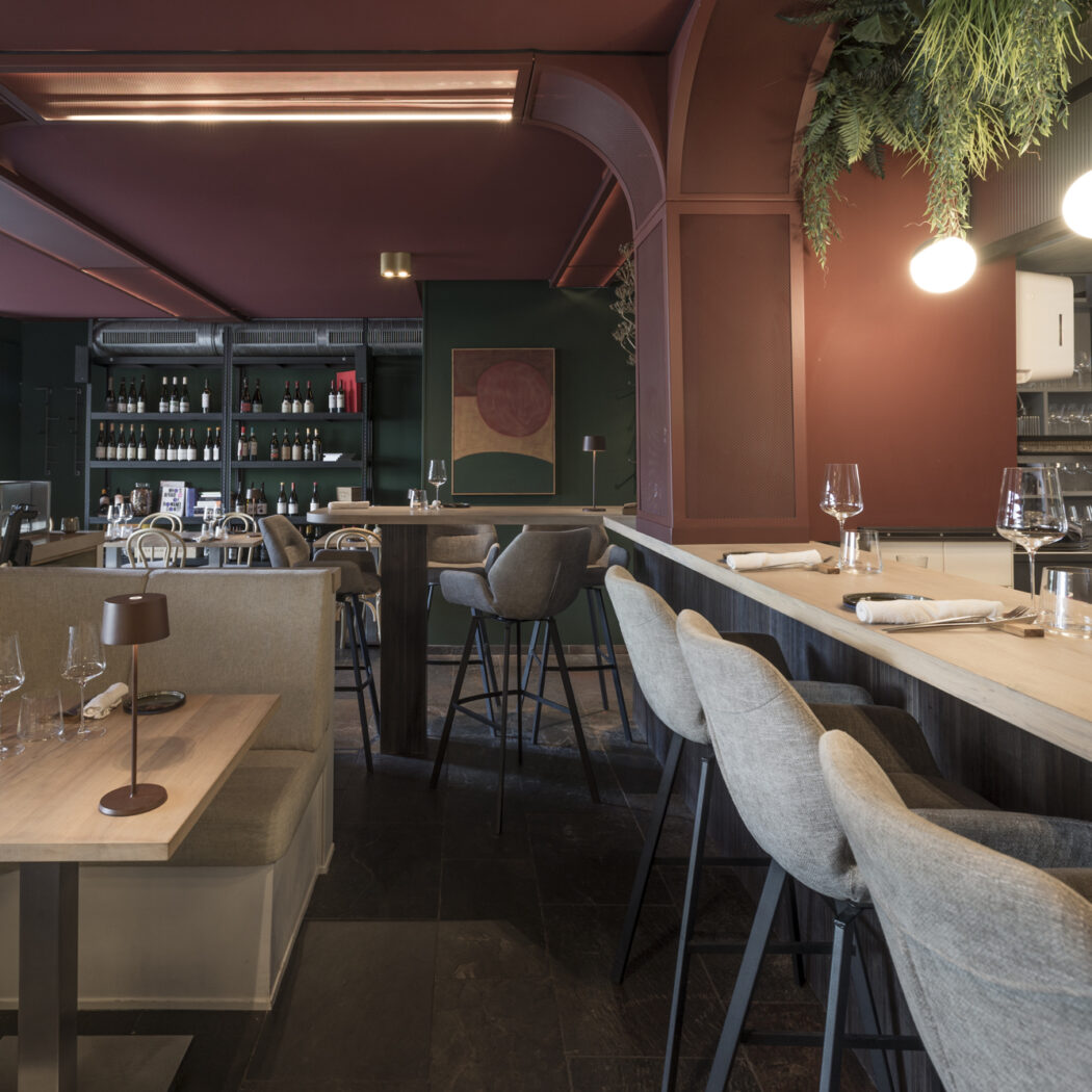 Modern restaurant met bordeauxrode muren, houten tafels, lichtgrijze barkrukken aan de bar en een open keuken.