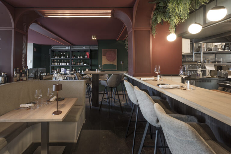 Modern restaurant met bordeauxrode muren, houten tafels, lichtgrijze barkrukken aan de bar en een open keuken.