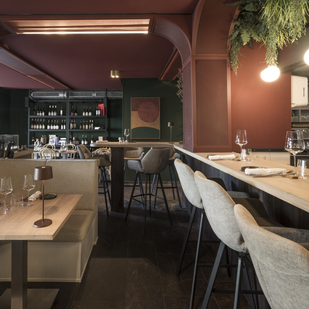 Modern restaurantinterieur met bankzitplaatsen, houten bar met barkrukken, rode en donkergroene muren en wijnflessen op planken.