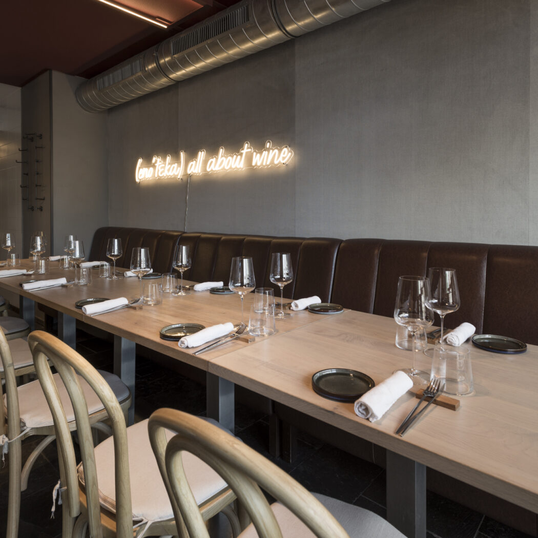 Modern restaurant interieur met een lange, gedekte houten tafel, bruine leren bank en neonreclame all about wine.