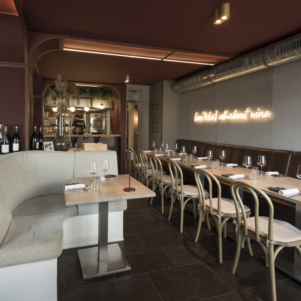 Modern restaurantinterieur met bordeauxrode plafonds, grijze muur met all about wine neonreclame, en diverse houten tafels en zitplaatsen.