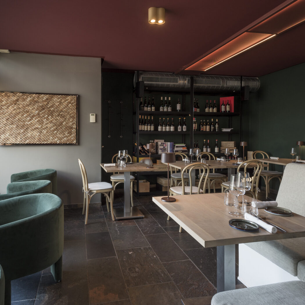 Sfeervol restaurant met groene fauteuils, houten tafels gedekt voor gasten, en een wijnrek tegen een donkergroene muur.