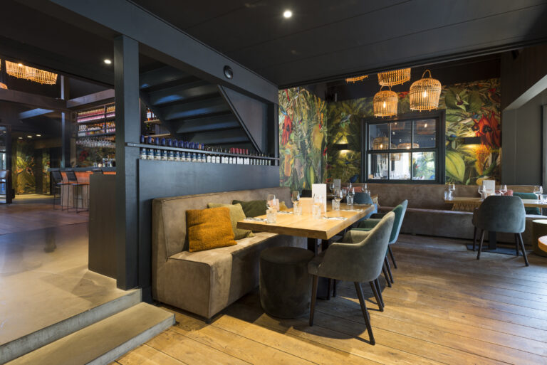 Restaurantruimte met banken, houten tafels, groen-rood tropisch behang, rieten lampen en een achterliggende bar.
