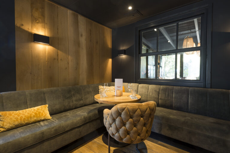 Gezellige zithoek in een restaurant met donkergrijze hoekbank, ronde houten tafel met MENU DRANKJES, gecapitonneerde stoel en raam.
