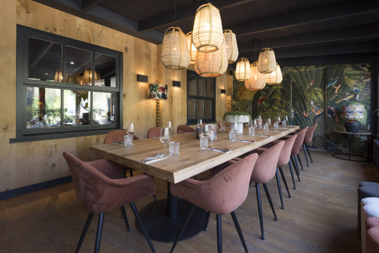 Lange gedekte houten tafel met roze fluwelen stoelen, rieten hanglampen en jungle behang in een sfeervolle eetruimte.