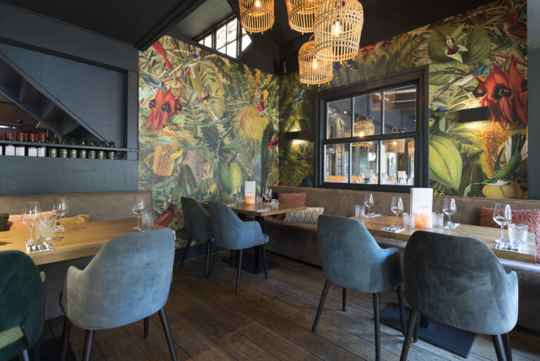 Sfeervol restaurant met weelderig tropisch behang, houten tafels, blauwe fluwelen stoelen en rotan lampen.