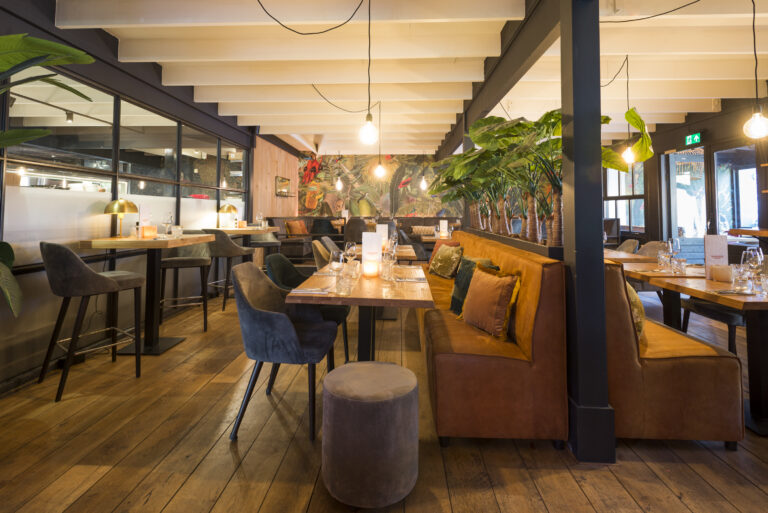 Sfeervol restaurant met houten vloer, bruine zitbanken, barstoelen en tropische planten bij kleurrijk behang.