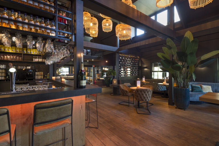 Sfeervol bar-restaurant interieur met donkere houten bar, champagnefles Moët & Chandon, rieten hanglampen en comfortabele zitjes.