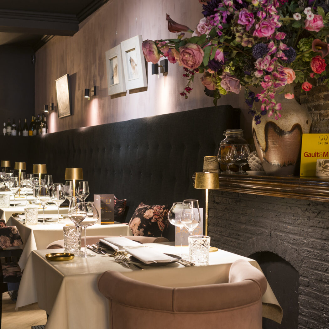 Elegant restaurant met gedekte tafels, donkere banquette en een sfeervolle schouw met bloemstuk en Gault&Millau bord.