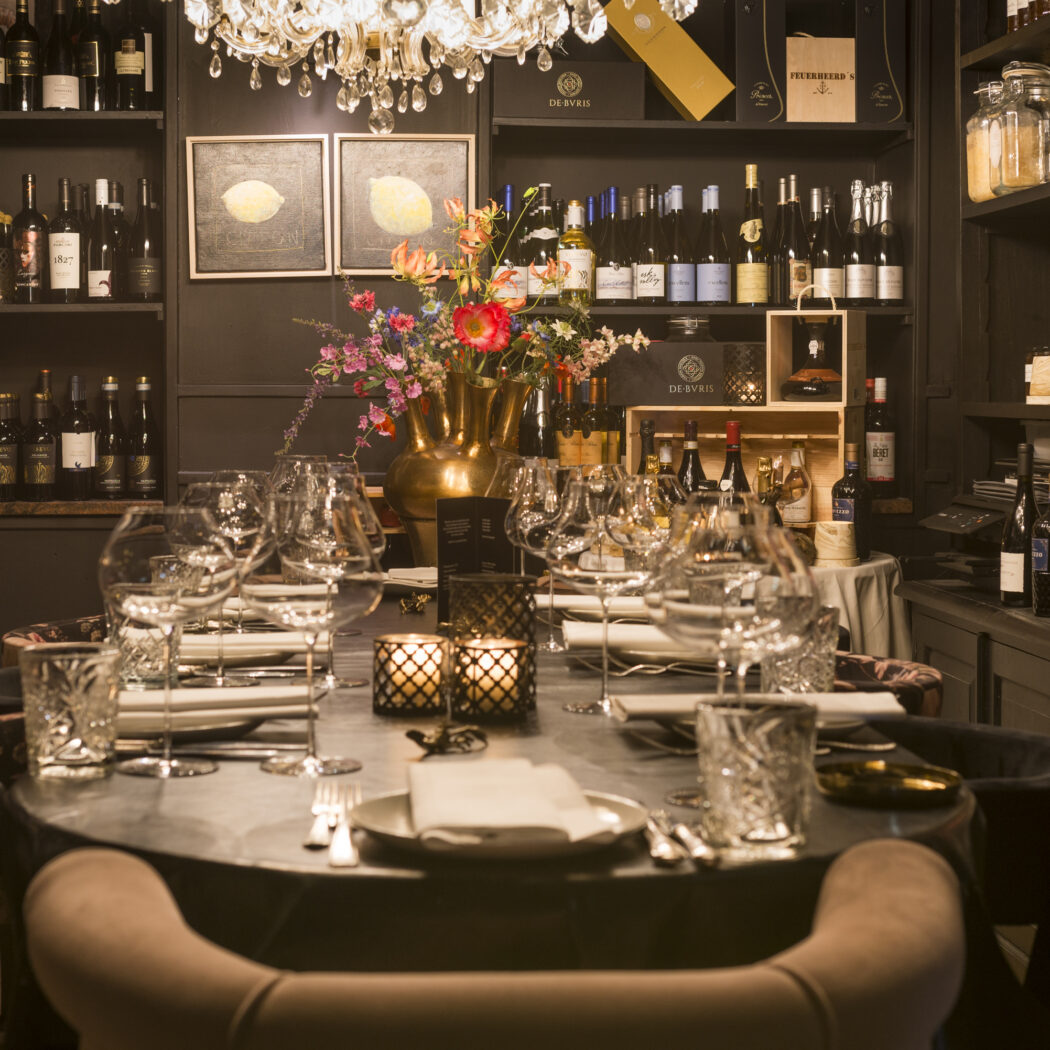 Gedekte ronde tafel met glazen, kaarsen en een bloemstuk onder een kroonluchter in een luxe wijnbar.
