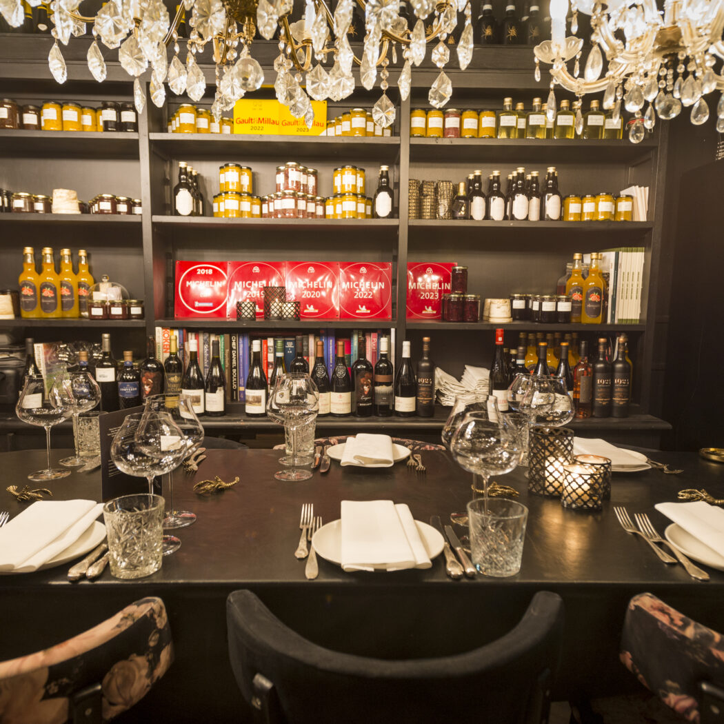Gedekte dinertafel onder kristallen kroonluchter, met planken vol Michelin awards, gidsen en delicatessen.