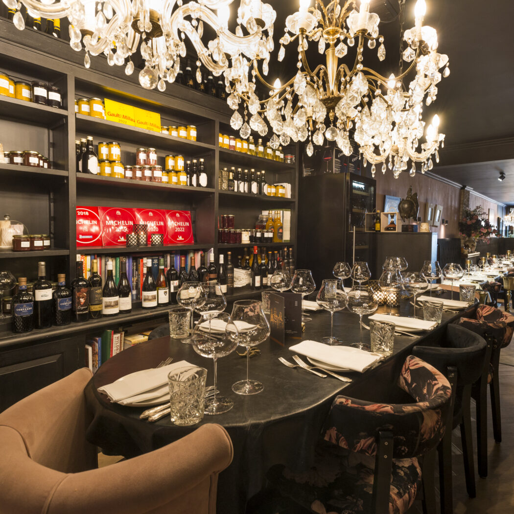 Elegant restaurantinterieur met kristallen kroonluchters, lange gedekte tafel en planken met wijnen en Michelin gidsen.