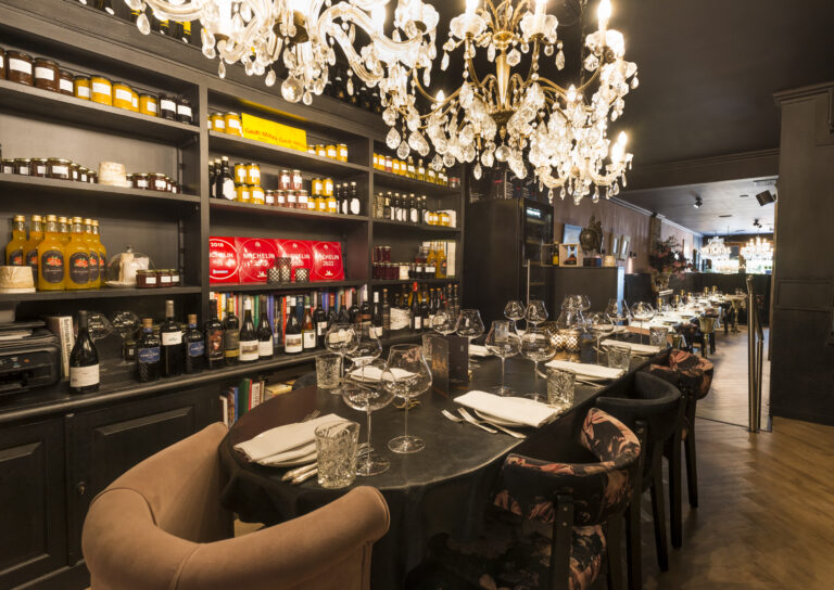 Elegant restaurantinterieur met kristallen kroonluchters, lange gedekte tafel en planken met wijnen en Michelin gidsen.