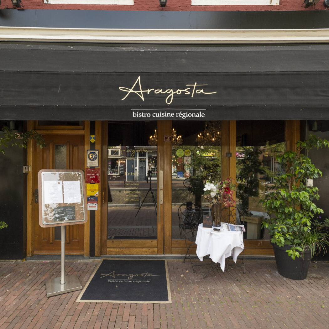 Voorgevel van Aragosta bistro cuisine régionale met zwarte luifel, houten deuren, planten en menukaart.