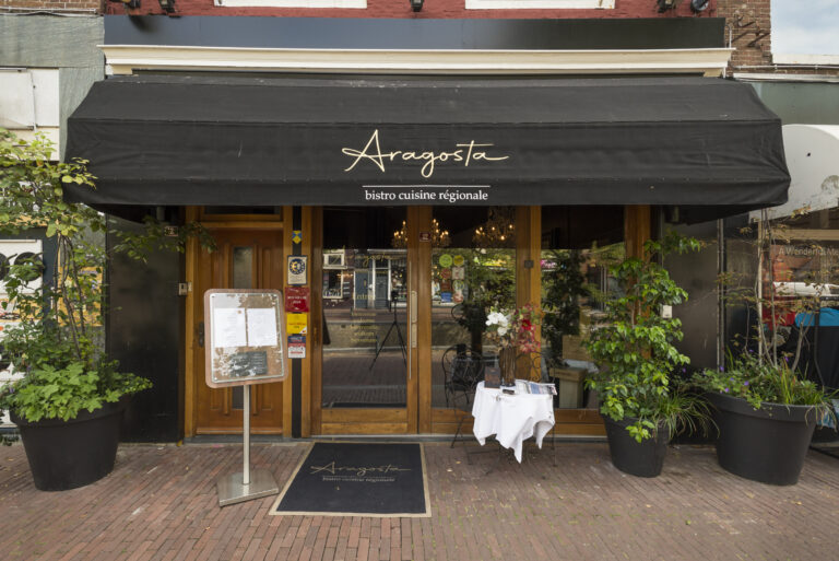 Voorgevel van Aragosta bistro cuisine régionale met zwarte luifel, houten deuren, planten en menukaart.