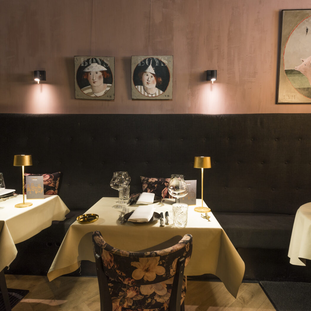 Sfeervol restaurant met roze muren, donkere bank, gedekte tafels met gouden lampen en opvallende schilderijen.