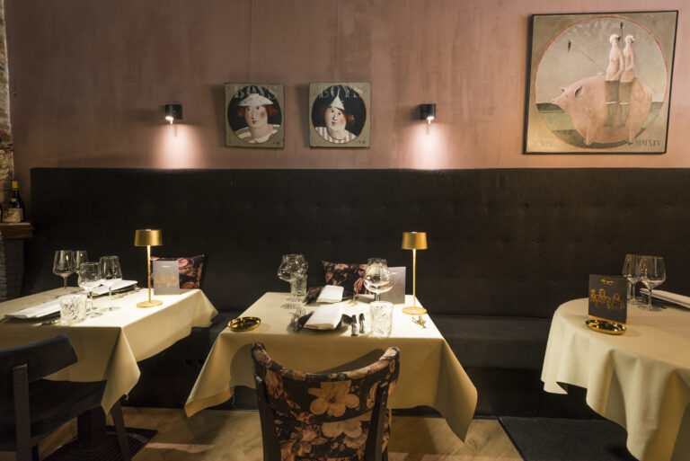 Sfeervol restaurant met roze muren, donkere bank, gedekte tafels met gouden lampen en opvallende schilderijen.