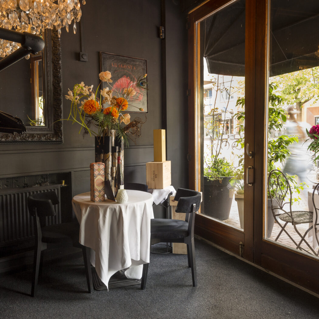 Sfeervol restaurantinterieur met spiegel, ronde tafel, oranje bloemen en glazen deuren naar terras.