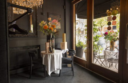Donker interieur met tafeltje, stoelen en bloemen, zicht op straat door glazen deur.