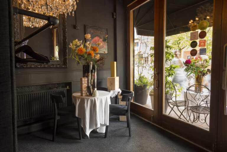 Sfeervol restaurantinterieur met spiegel, ronde tafel, oranje bloemen en glazen deuren naar terras.