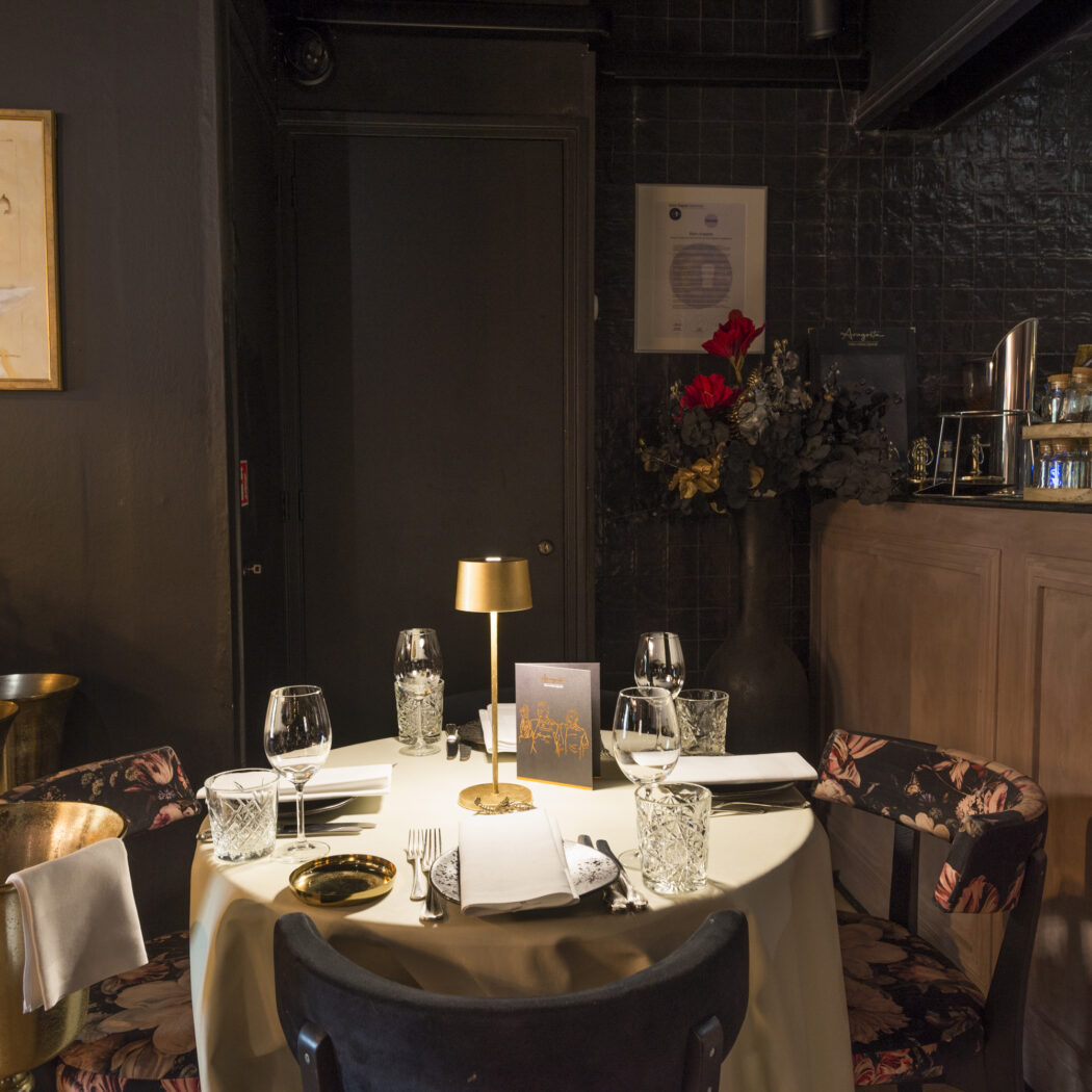 Gedekte ronde tafel met gouden tafellamp, bloemstuk en bar in een donker, sfeervol restaurantinterieur.