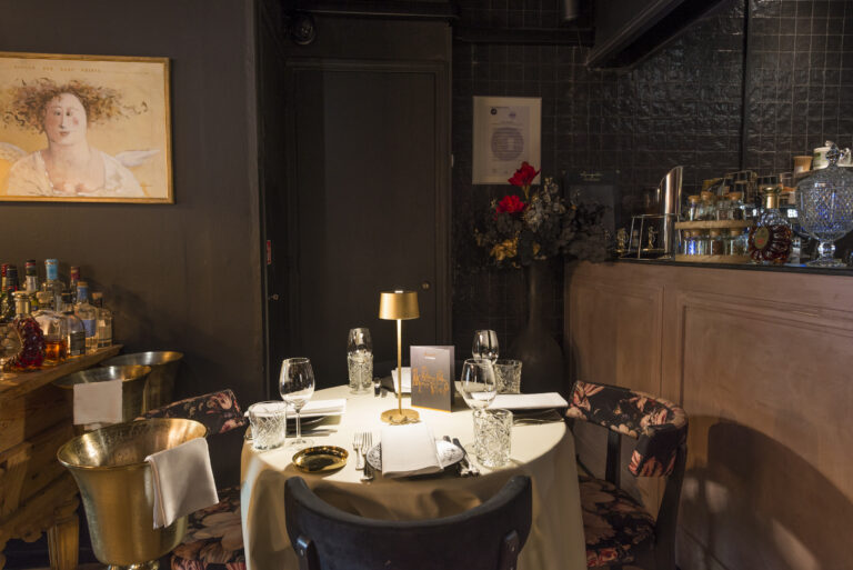 Gedekte ronde tafel met gouden tafellamp, bloemstuk en bar in een donker, sfeervol restaurantinterieur.
