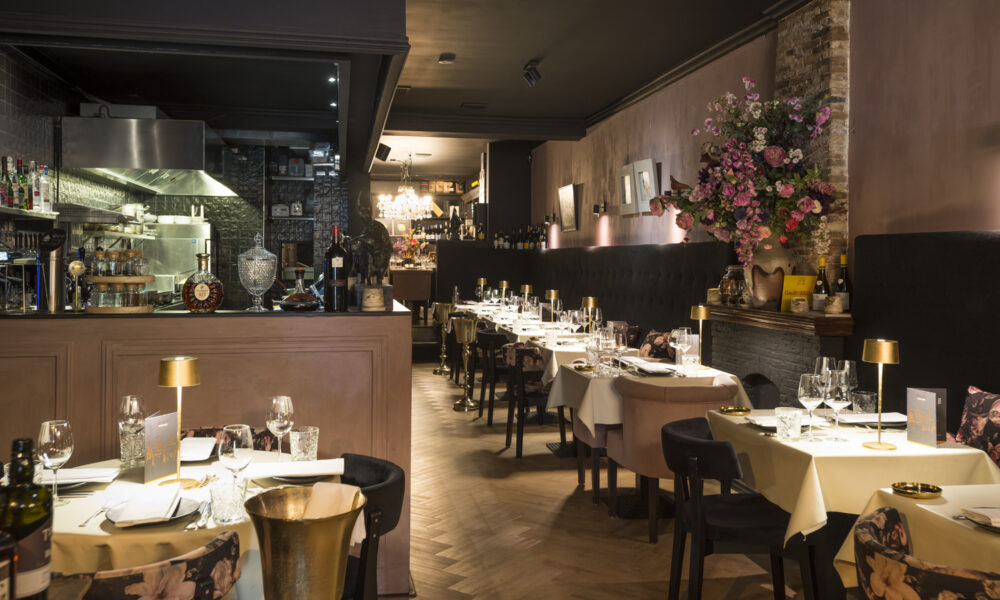 Restaurant interieur: gedekte tafels, visgraatvloer, bar met keuken zichtbaar.