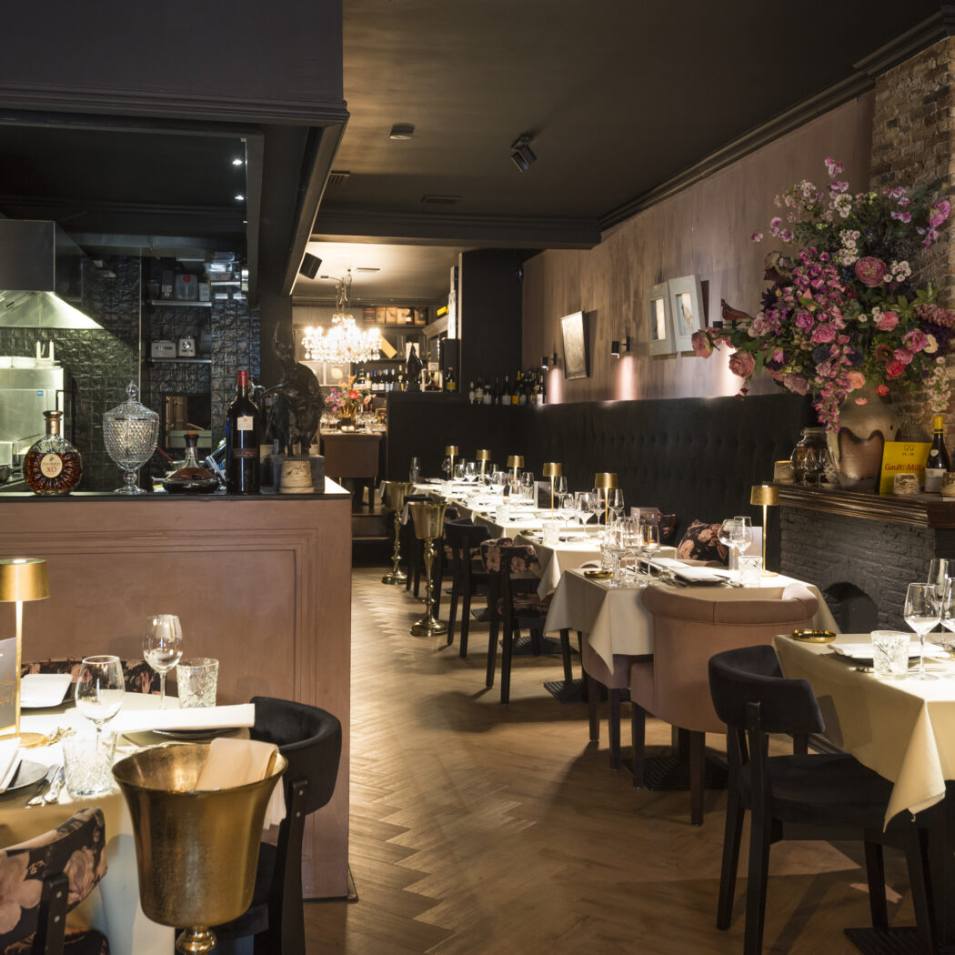 Sfeervol restaurantinterieur met gedekte tafels, een open keuken en een groot bloemenarrangement.