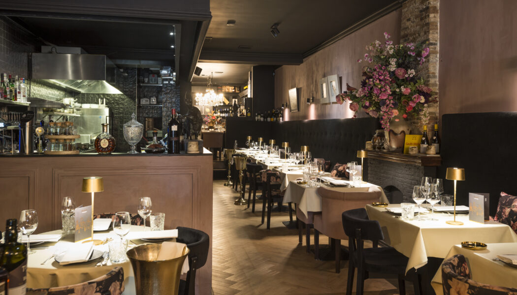 Restaurant interieur: gedekte tafels, visgraatvloer, bar met keuken zichtbaar.