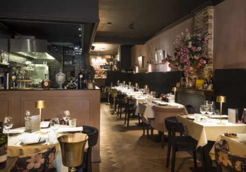 Sfeervol restaurantinterieur met gedekte tafels, een open keuken en een groot bloemenarrangement.