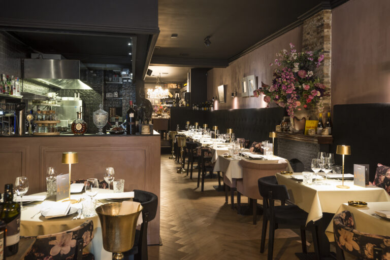 Sfeervol restaurantinterieur met gedekte tafels, een open keuken en een groot bloemenarrangement.