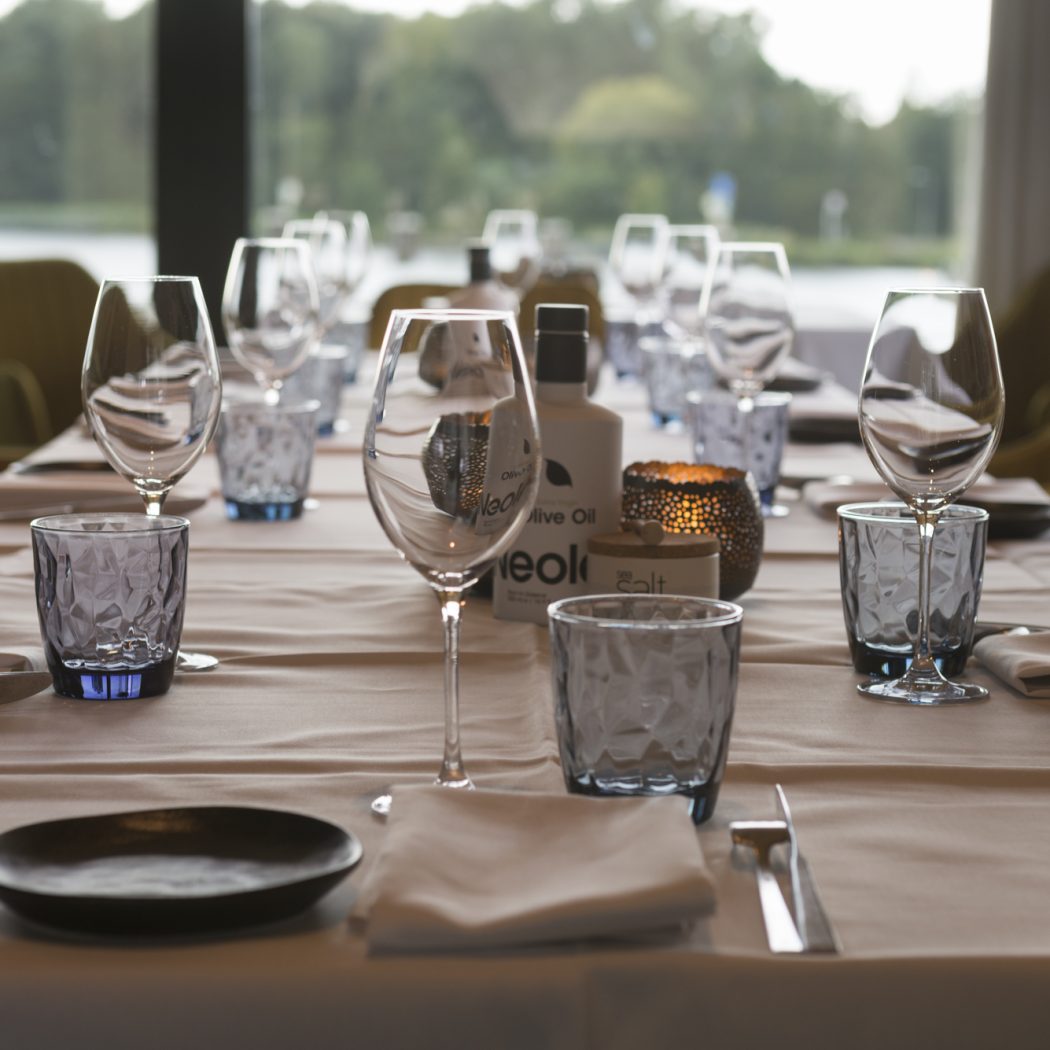 Elegant gedekte restauranttafel met wijnglazen, blauw glaswerk, olijfolie en kaarslicht, tegen een raam met waterzicht.