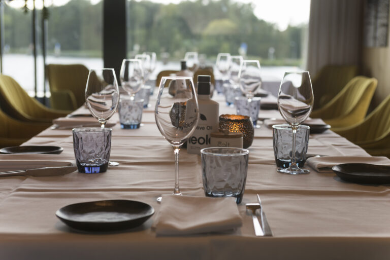 Elegant gedekte restauranttafel met wijnglazen, blauw glaswerk, olijfolie en kaarslicht, tegen een raam met waterzicht.