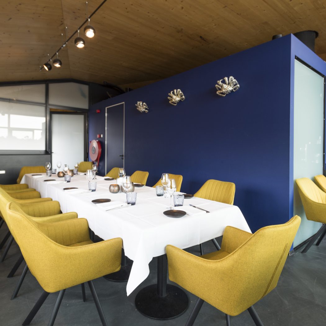 Moderne eetruimte met lange gedekte tafel, gele stoelen, blauwe muur met zilveren decoratie en houten plafond.
