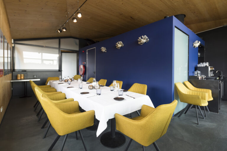 Moderne eetruimte met lange gedekte tafel, gele stoelen, blauwe muur met zilveren decoratie en houten plafond.
