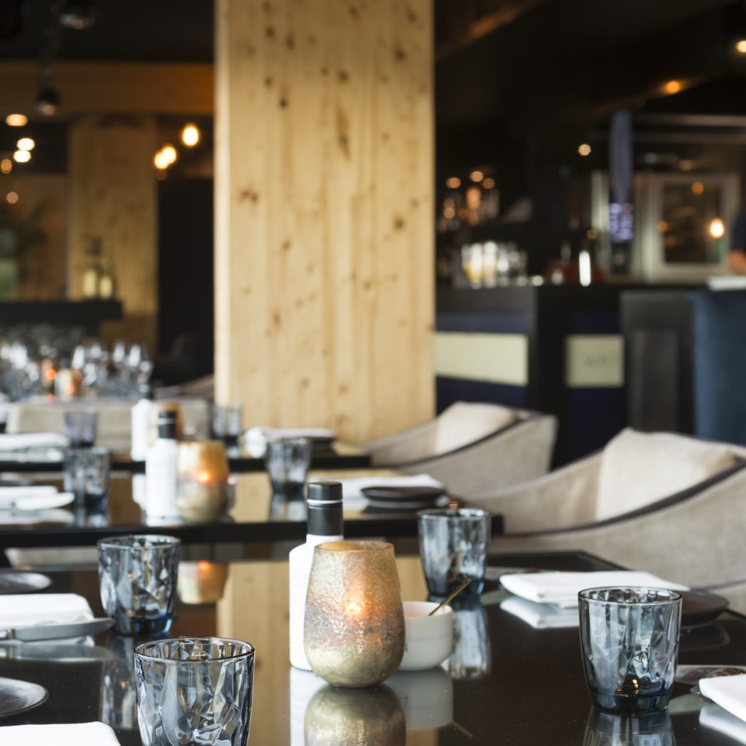 Een moderne restaurantzaal met gedekte zwarte tafels, facetglazen, witte servetten en brandende kaarsen.