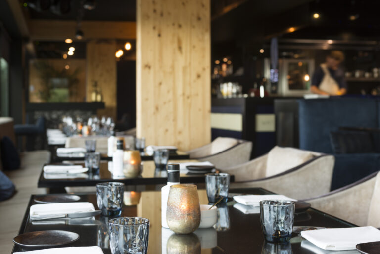 Een moderne restaurantzaal met gedekte zwarte tafels, facetglazen, witte servetten en brandende kaarsen.