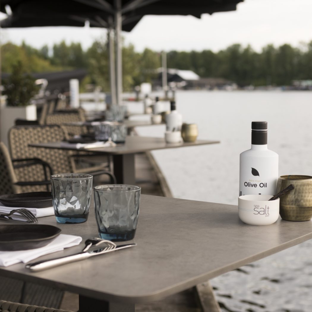 Een gedekte restauranttafel op een terras aan het water met een fles olijfolie, zeezout en twee glazen.