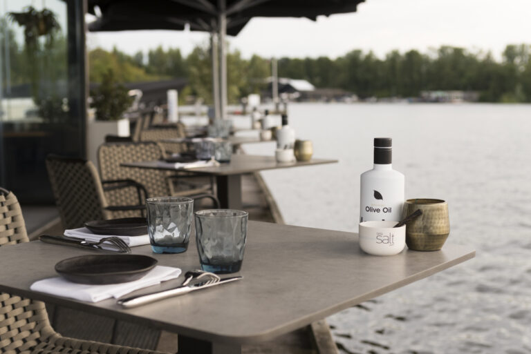 Een gedekte restauranttafel op een terras aan het water met een fles olijfolie, zeezout en twee glazen.