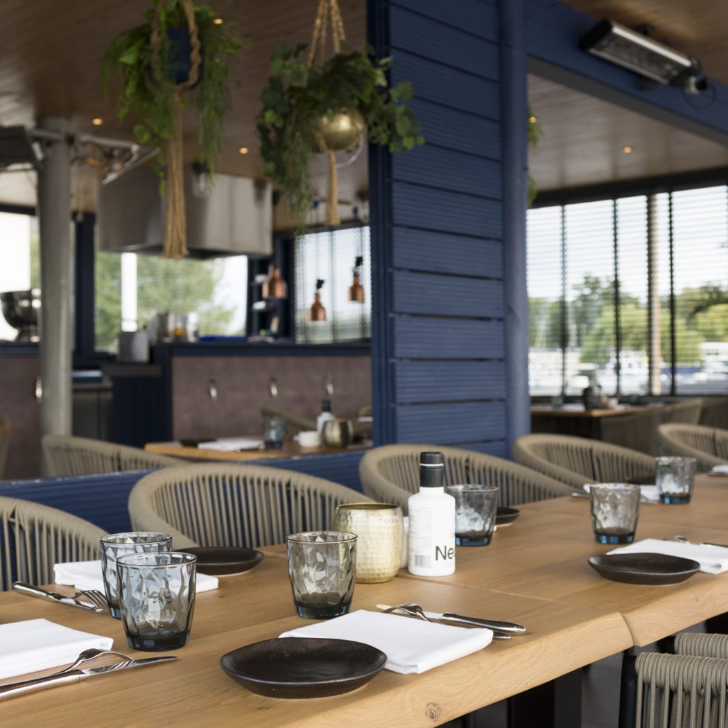 Lange houten tafel met donkere borden, glazen en rieten stoelen op een modern restaurantterras met blauwe muren en planten.