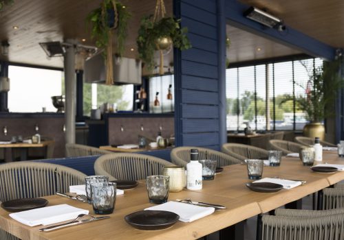 Lange houten tafel met donkere borden, glazen en rieten stoelen op een modern restaurantterras met blauwe muren en planten.