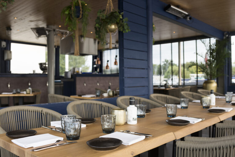 Lange houten tafel met donkere borden, glazen en rieten stoelen op een modern restaurantterras met blauwe muren en planten.