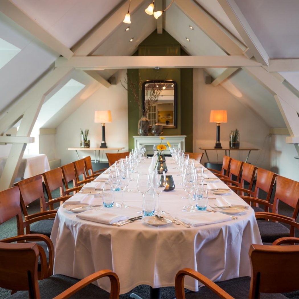 Lange, elegant gedekte private dining tafel met houten stoelen onder een schuin plafond met balken.